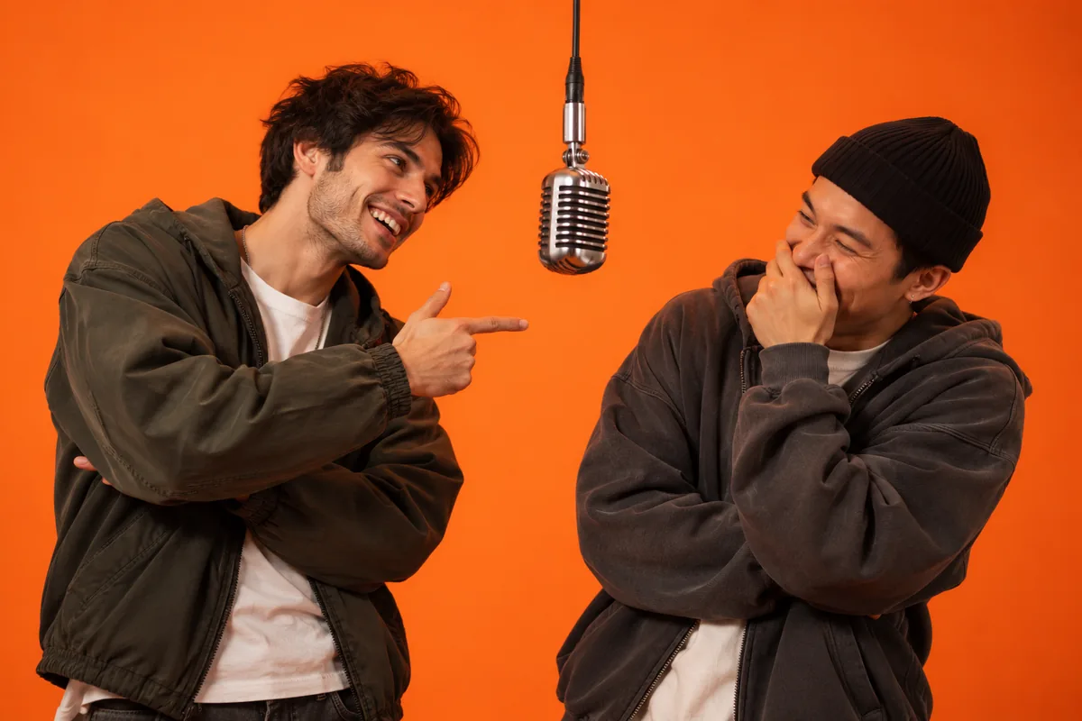 Two friends at a hanging studio mic, one pointing with a teasing grin while the other laughs behind his hand