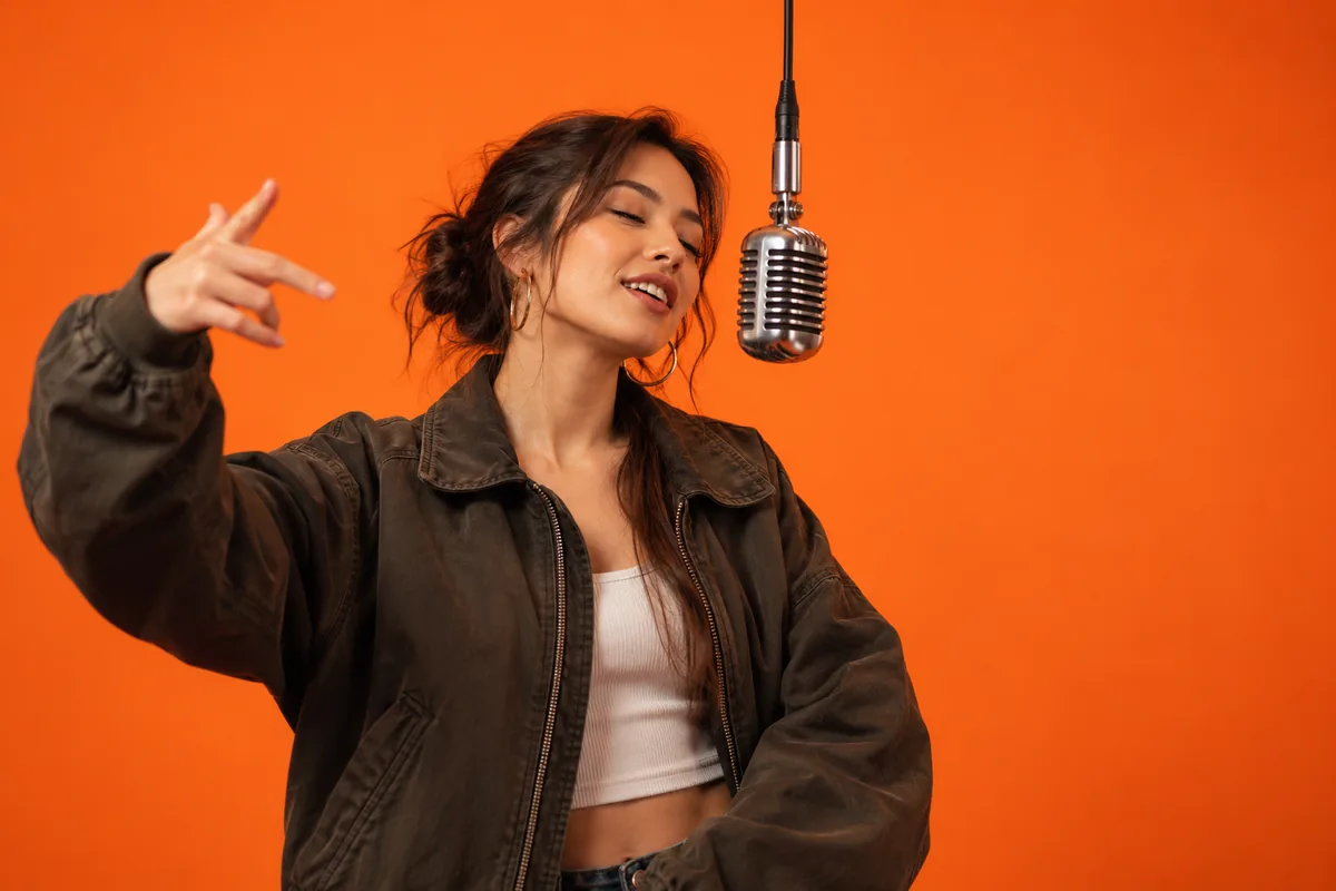 A young woman alone at a hanging studio mic in front of an orange wall, one hand raised mid-verse